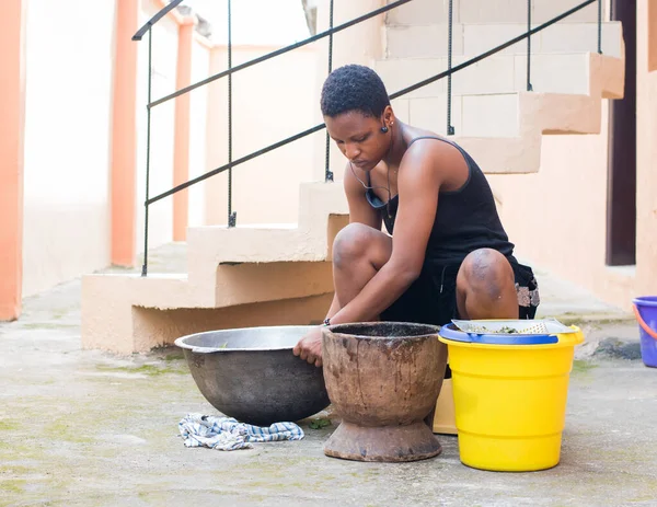 An African young girl, lady or woman from Nigeria doing house chores like cooking and working with a big pot, wooden mortar and bucket to prepare edible vegetables for food, herb or medications