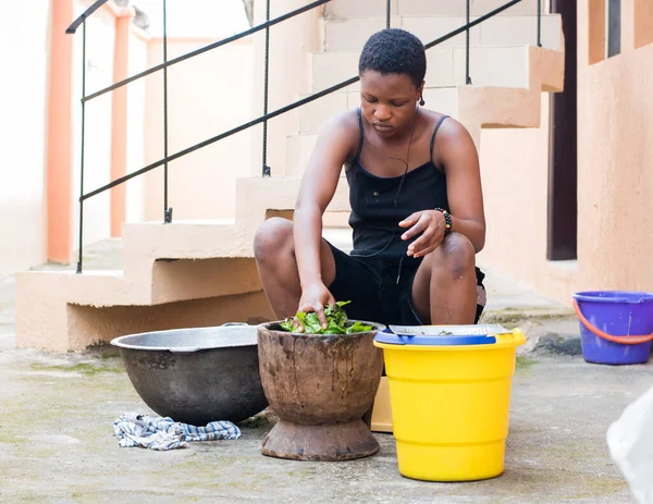 An African young girl, lady or woman from Nigeria using a wooden pestle and mortar to pound and grind edible vegetables for food cooking, herb or medications