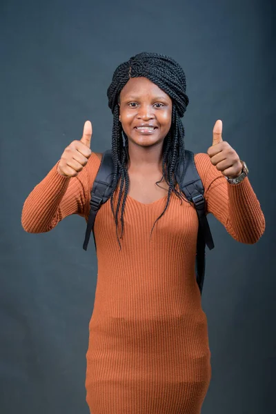 A happy African lady, female or student from Nigeria happily doing thumbs up gestures with both her hands with a black school bag on her back