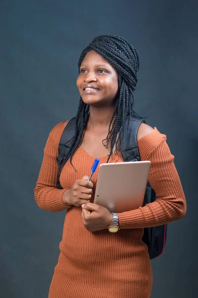 A happy African lady, female or student from Nigeria happily looking at the camera while carrying an education smart tablet in her hands and a school bag on her back