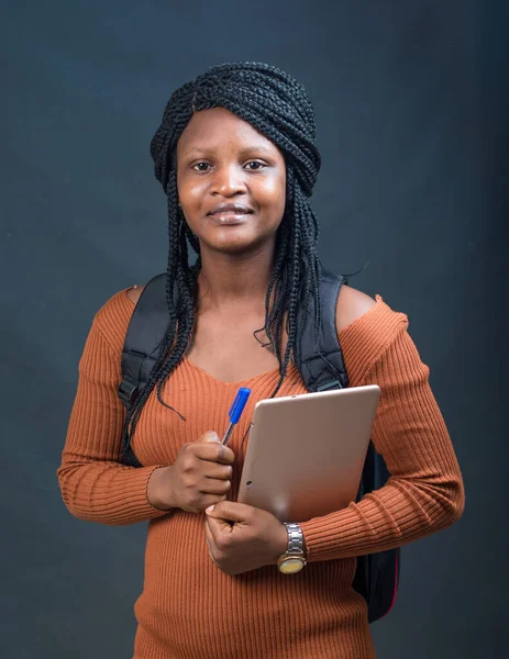 A happy African lady, female or student from Nigeria happily looking at the camera while carrying an education smart tablet in her hands and a school back on her back