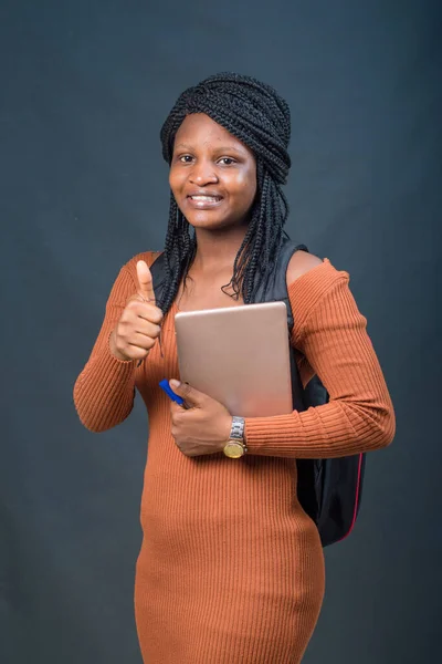 A happy African lady or female student from Nigeria happily doing thumbs up to the camera while carrying an education smart tablet in her hands and a school back on her back