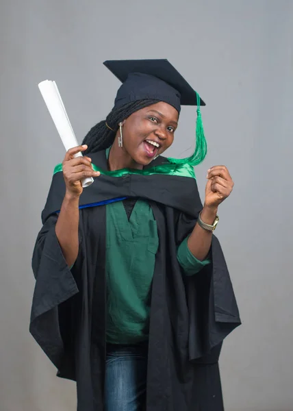 Portrait of an excited African female student or graduand from Nigeria, wearing graduation gown and cap while posing for the camera and celebrating success in education