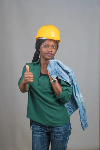A cute African lady or woman from Nigeria with yellow safety helmet, doing thumbs up gestures as she represents industrial professionals like construction specialist, builders, architect and engineer