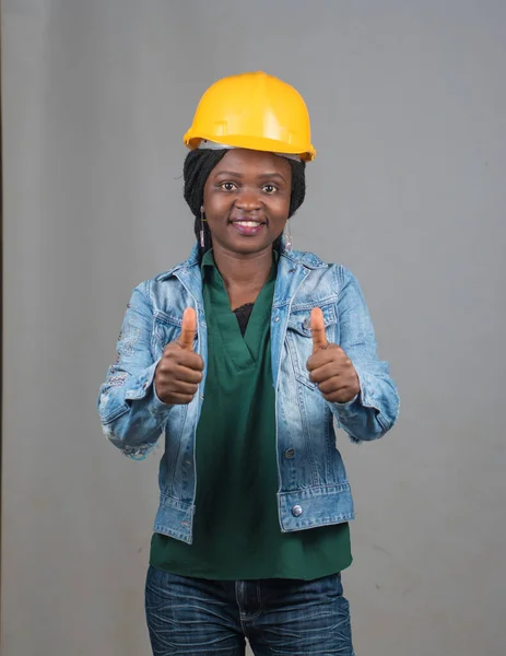 Happy African lady or woman from Nigeria with yellow safety helmet, doing thumbs up gestures as she represents industrial professionals like construction specialist, builders, architect and engineer
