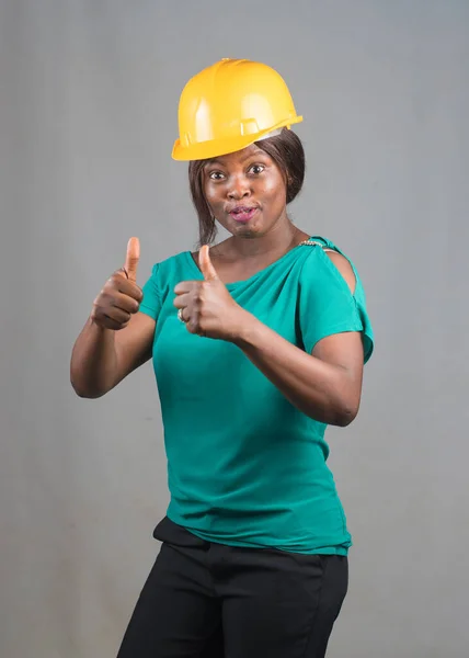 Happy African lady or woman from Nigeria with yellow safety helmet, doing thumbs up gestures as she represents industrial professionals like construction specialist, builders, architect and engineer