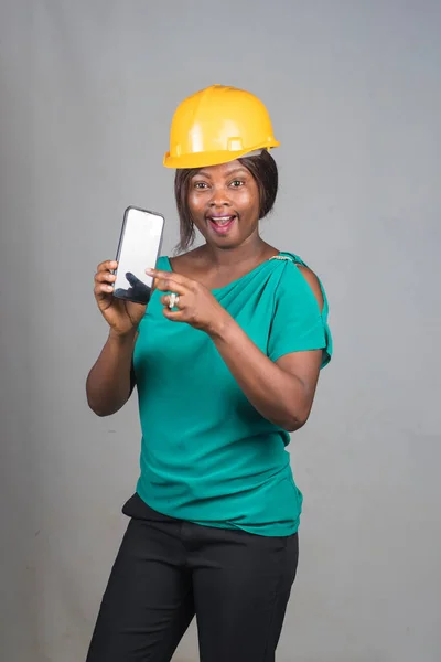 A cute African lady or woman from Nigeria with yellow safety helmet pointing to a smart phone while representing industrial professionals like construction specialist, builders, architect and engineer