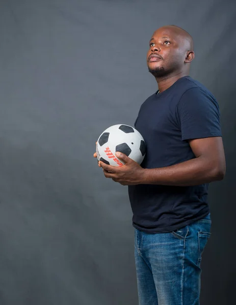 A studio portrait of a happy African man or guy holding a black and white football to his body while looking at something up ahead