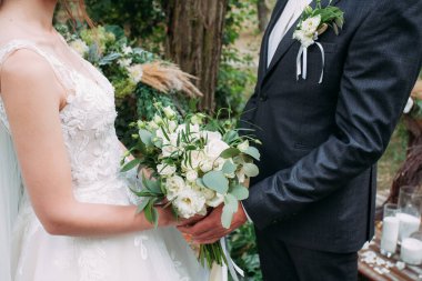 Yeni evliler doğadaki bir düğün töreninde bir buket beyaz gülle birbirlerinin elini tutarlar. Gelin güzel beyaz bir elbise, damat koyu mavi bir takım ve yaka çiçekli bir elbise giyiyor..