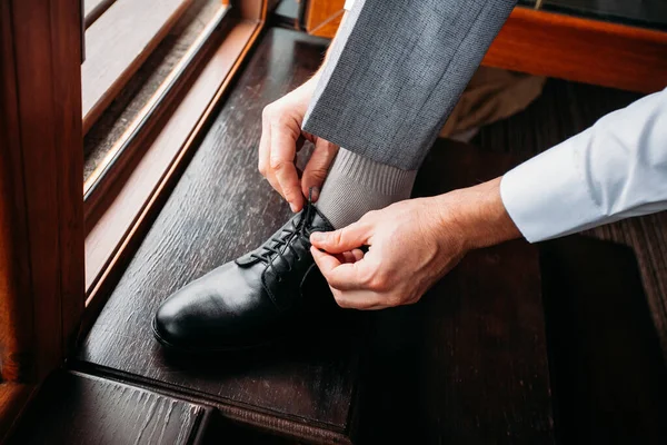 Damat ayakkabı bağcıklarını siyah şık ayakkabılara bağlıyor. Sabahları, kapalı bir yerde, ahşap kahverengi bir zeminde.