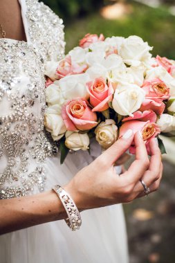 Gümüş takılar takmış şık beyaz gelinlik içinde genç bir gelin elinde taze pembe ve sarı güllerden bir buket tutuyor ve parmaklarıyla nazikçe ona dokunuyor..