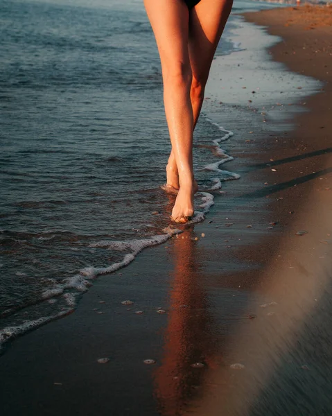 Beautiful athletic legs of a girl walk along the waves near the seashore. Image with reflection in water.