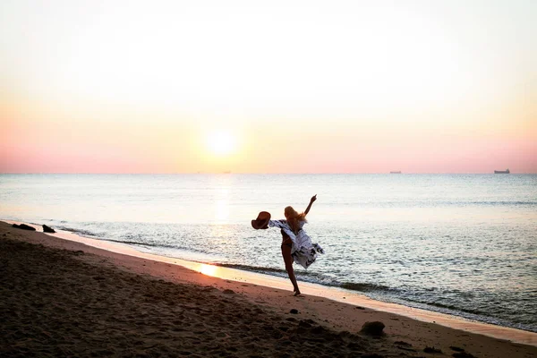 Art work silhouette of a happy girl with a hat, running along the sandy shore of the ocean, near the hill, in the morning at dawn. The lady raises her hands.