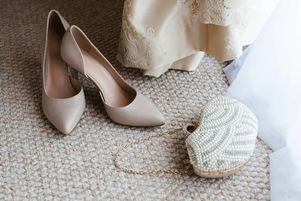 Fashionable women\'s accessories lie on the floor in the room. Beige shoes and a bride\'s handbag, a cream-colored wedding dress hangs nearby.