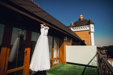 Modaya uygun beyaz gelinlik otelin penceresinden sarkan mavi gökyüzüne karşı. Gelinin sabahının ayrıntıları. Yerde yeşil halı var..