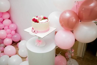 Canlı kırmızı, beyaz ve pembe çiçeklerle süslenmiş iştah açıcı sarı bir pasta pembe ve beyaz balonlarla süslenmiş bir odada beyaz bir dekoratif ayakta duruyor..