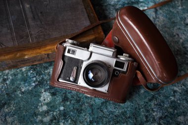 Kahverengi deri bir çantanın içindeki eski bir Sovyet kamerası desenli mavi bir masanın üzerinde duruyor. Yakınlarda eski bir kitap var..