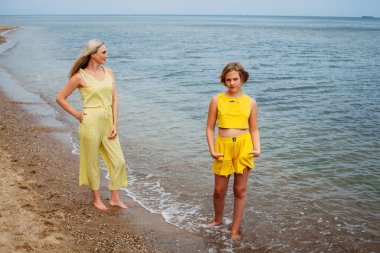 Sarı elbiseli güzel anne ve kız güneşli bir günde kumlu sahilde duruyor..