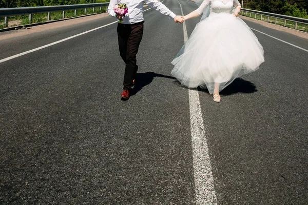 花嫁と新郎は舗装された道路に沿って手をつないで走ります 黒いズボンと白いシャツの新郎は彼の手に花束を持っています 花嫁は壮大な白いドレスを着ています — ストック写真