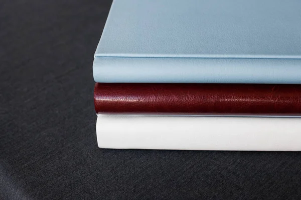Three stylish photobooks with leather covers, white, burgundy and blue, of different thicknesses, lie on a dark gray surface in a room.