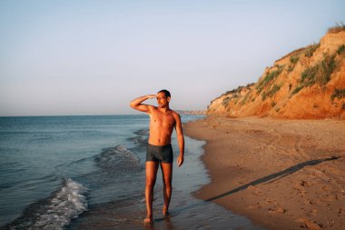 Mavi şortlu, ince, bronz tenli bir adam okyanusun kumlu kıyılarında yürür ve parlak güneşli bir günde mesafeye bakar. Tepeler uzaktan görülebilir..