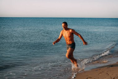 Mavi yüzme şortlu ince kaslı bir adam okyanusun kumlu kıyılarında koşuyor ve güneşli bir günde gülümsüyor..