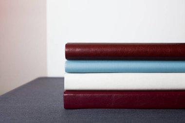 Deri kapaklı dört şık fotoğraf kitabı, beyaz, bordo, kahverengi ve mavi, farklı kalınlıkta, gri bir masanın üstünde, açık bir duvara yaslanmış,.