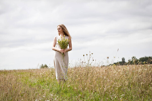 A slender girl in a linen summer dress, with blond long hair and a bouquet of wild flowers in her hands, stands in a clearing during the day.