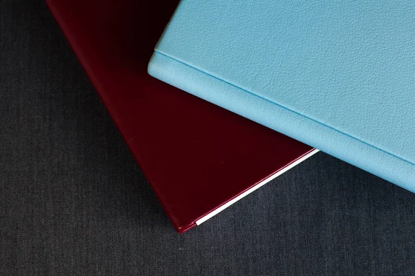 Deri kaplamalı iki şık fotobook, bordo ve mavi, farklı kalınlıkta, bir odada koyu gri bir yüzey üzerinde uzanır..