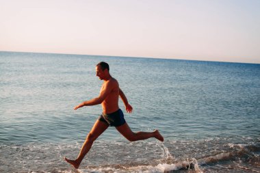 Mavi yüzme şortlu ince kaslı bir adam okyanusun kumlu kıyılarında koşuyor ve güneşli bir günde gülümsüyor..