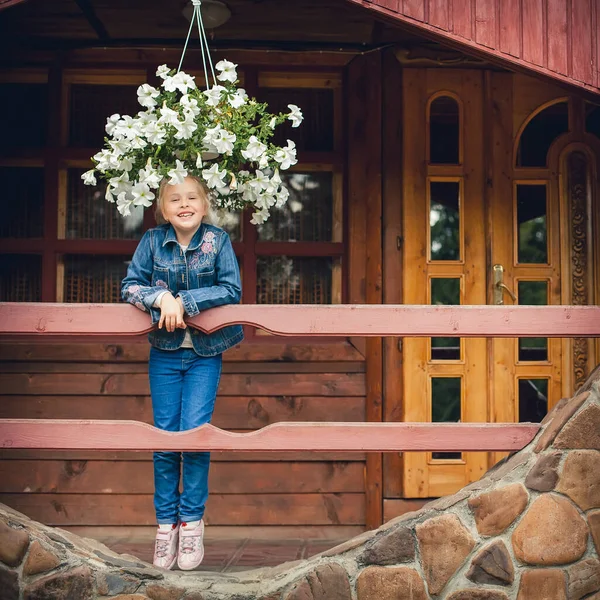 Little cute girl with blond hair laughs. Dressed in a denim suit, she stands at the wooden railing on the veranda of the cottage, decorated with flowers and vegetation.