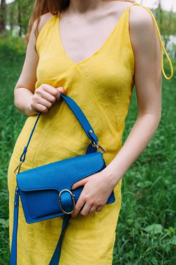 Sarı elbiseli zayıf bir kız çimenlerin arkasında mavi deri bir çanta tutuyor..