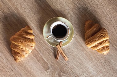 Bir fincan sıcak kokulu siyah kahve ayçöreği ve tarçın çubuklarıyla bir fincan çay tabağında, açık kahverengi ahşap bir masada..