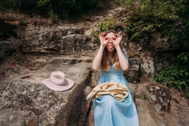 Mavi elbiseli genç bir kız güneşli bir günde dağlardaki kayalıklarda oturur ve yüzünü buruşturur. Yakınlarda pembe bir şapka ve bej bir çanta var..