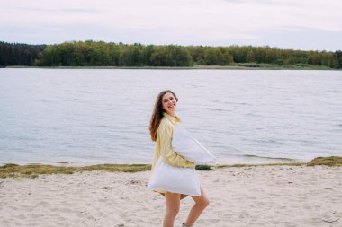 Sarı kıvırcık saçlı bir kız, sarı gömlekli, elinde bir sepet ve yastıkla, nehir kıyısında kumsalda yürüyor ve gülümsüyor..