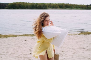 Sarı kıvırcık saçlı, sarı gömlekli, elinde sepet ve yastıkla nehir kıyısında kumsalda yürüyüp gülümseyen bir kız. Sepette bir şişe şampanya var..