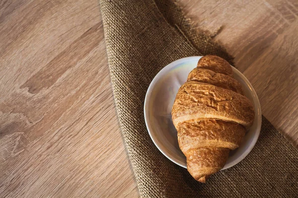 An appetizing croissant lies in a saucer on a fabric, on a wooden light brown textured table.