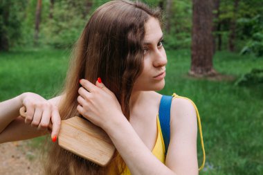 Sarı elbiseli, mavi deri çantalı, ince bir kız parkta duruyor ve uzun kıvırcık saçlarını tarıyor..