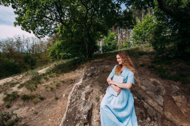 Genç bir kız doğada dizüstü bilgisayarla çalışıyor. Mavi bir yaz elbisesi giymişti. Ormanda taşların üzerinde oturuyor..