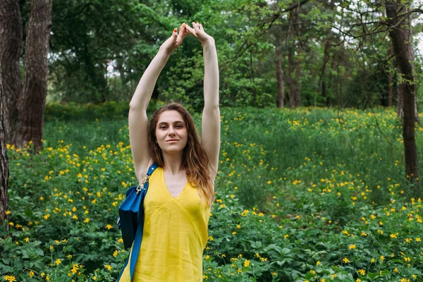 A young girl with blond curly hair in a yellow summer dress and a blue handbag, stands on a flowering lawn in the forest, raise your hands up and laughs.