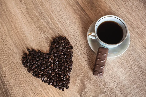 心臓の形で散乱コーヒー豆の背景に 木製の明るい茶色のテーブルの上に ソーサーとチョコレートで熱い芳香のコーヒーのカップ — ストック写真