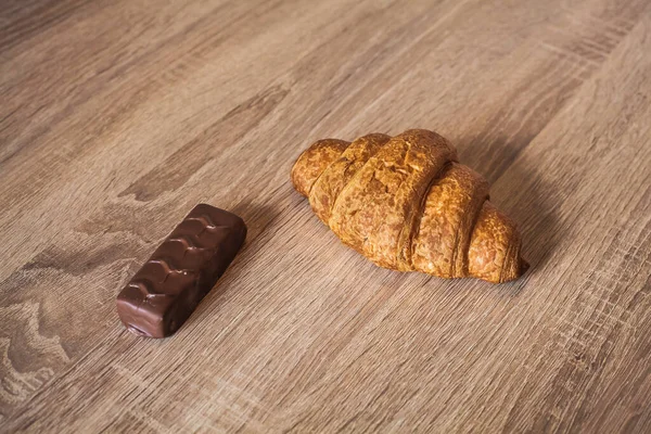 An appetizing croissant and a chocolate bar lie on a wooden light brown textured table, in the morning for breakfast.