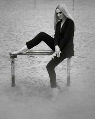 Sanat eserleri. Siyah beyaz bir fotoğraf, güzel bir stil kızı, sarı saçlı, siyah klasik takım elbiseli, plajdaki ahşap bir bankta oturuyor..