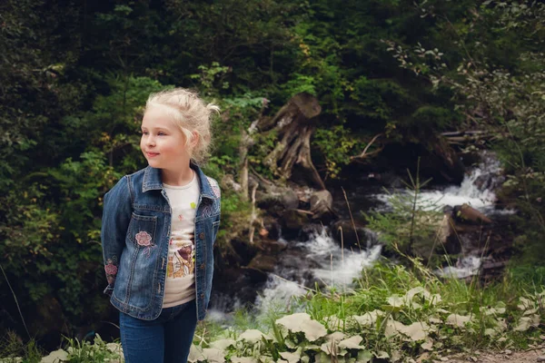 Sarı saçlı, kot elbiseli küçük bir kız yazın, taşlı ve ormanlı bir dağ nehrinin arka planında duruyor..