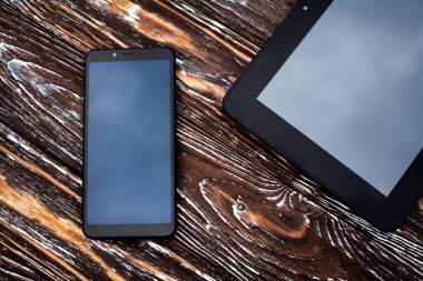 İki siyah cihaz, bir tablet ve bir cep telefonu koyu ahşap desenli bir yüzeye uzanıyor..