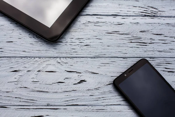 Elements of devices, a tablet and a mobile phone lie on a light wooden textured surface.