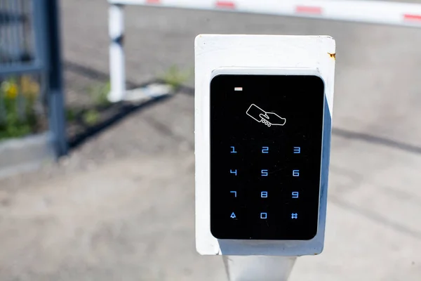 A card reader for access cards for entering the parking lot, in the background there is a white barrier with red stripes blocking the entrance.