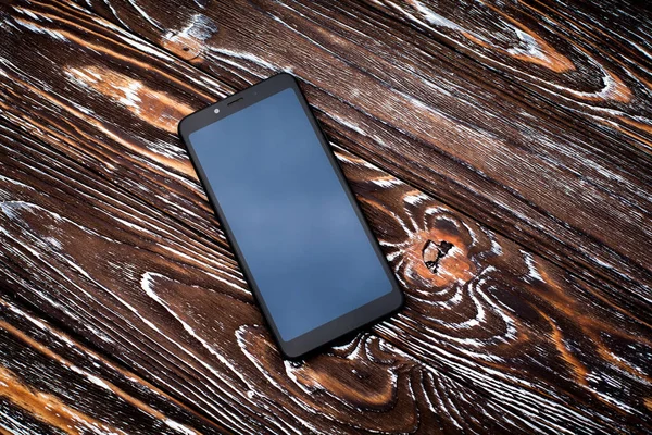 A black mobile phone with a dark display without an image lies on a dark wooden textured surface.