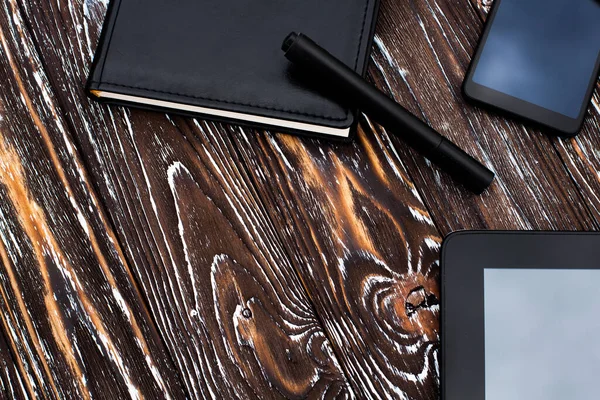 A black tablet, mobile phone and notebook with a leather cover and a pen lie on a dark wooden textured surface.