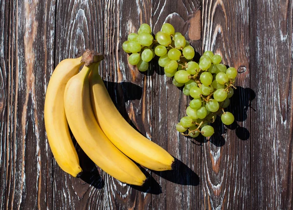 Still life of three ripe appetizing bananas and a bunch of grapes, on a dark brown wooden textured table, for your design or illustrations.